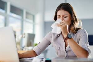 Una donna seduta al computer si soffia il naso per colpa dell'allergia