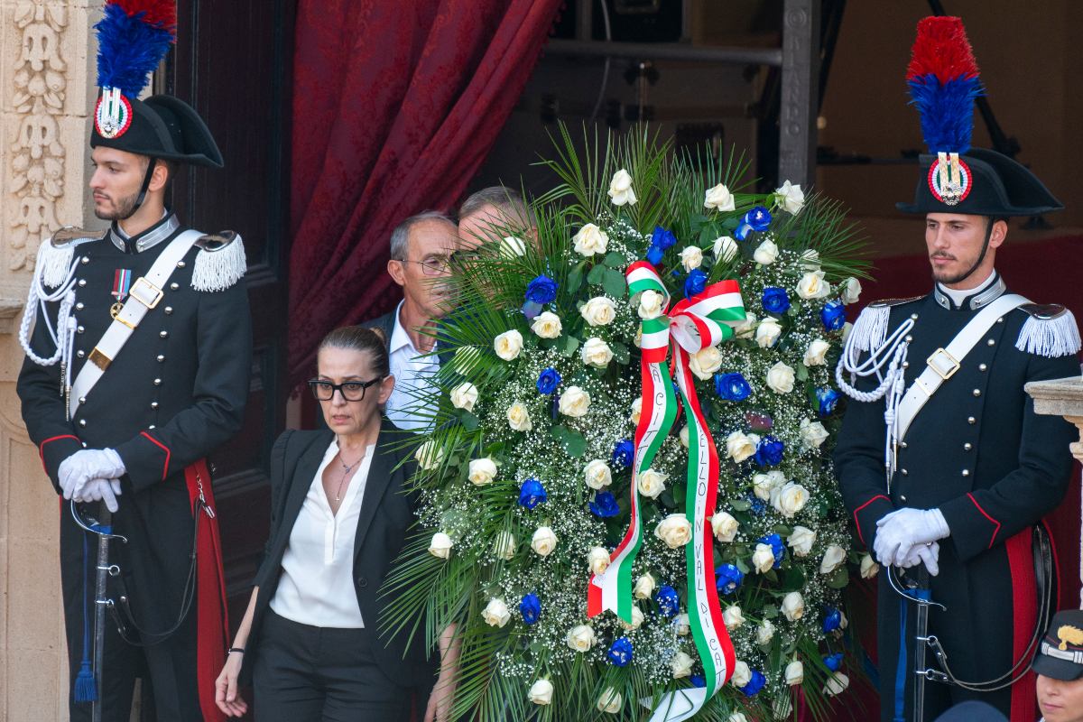 Dina Minna, storica segretaria del conduttore televisivo Pippo Baudo, con occhiali scuri e visibilmente commossa tra la folla, durante i funerali al Santuario di Santa Maria della Stella. Sullo sfondo, carabinieri in alta uniforme vigilano accanto a una corona di fiori deposta in omaggio, a Militello in Val di Catania, il 20 agosto 2025, in provincia di Catania.