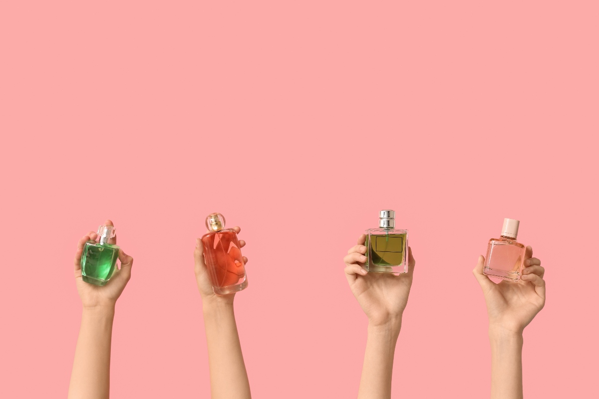 Sfondo rosa con mani di donne che sorreggono vari flaconi di profumo.