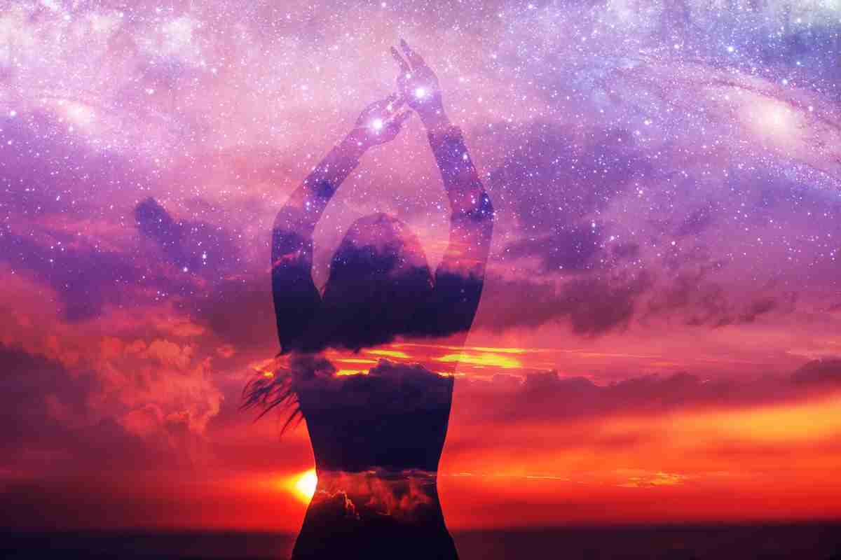 Sagoma di una giovane donna in piedi con le mani protese verso l’alto a toccare le stelle sulla riva del mare al tramonto, sullo sfondo dello spazio e del cielo stellato